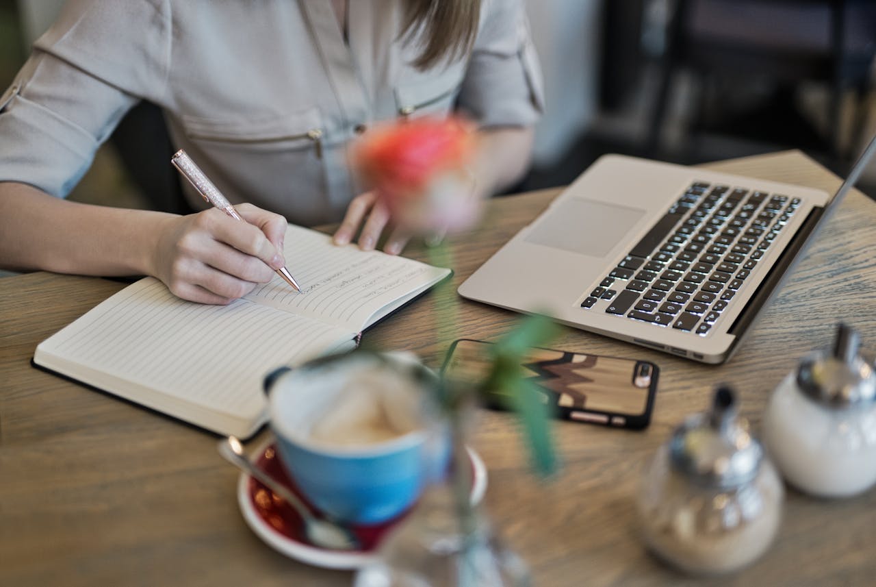 Home Woman writing in a notebook with a laptop and coffee cup on a desk. Ideal for workspace inspiration.